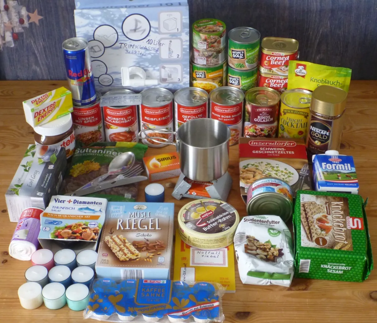 Ausgebreiteter Lebensmittelvorrat für eine Woche: Konserven, Trinkwasserbehälter, Knäckebrot, Müsliriegel, Kaffee, Teelichter, Esbit-Kocher mit Topf und Besteck auf einem Holztisch